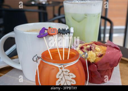 A festive Halloween cafe table setting featuring a mug of coffee, an iced matcha latte, and a berry crumble muffin arranged alongside a decorative fro Stock Photo