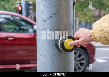 a pedestrian's hand pressing a round yellow crossing button on a metal pole while a red car passes by a red traffic signal in the background. Stock Photo