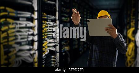 A technician wearing safety helmet holding tablet and stylus in a data center, checking server systems, network infrastructure and IT equipment mainte Stock Photo