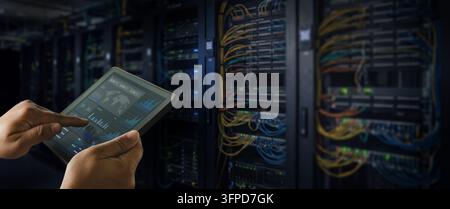 man or technician using tablet dashboard beside network and server racks, monitoring network systems and managing data center IT infrastructure. Stock Photo