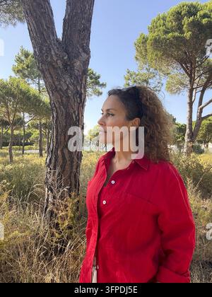 Woman in a red shirt standing thoughtfully among pine trees in a sunny Mediterranean woodland Stock Photo