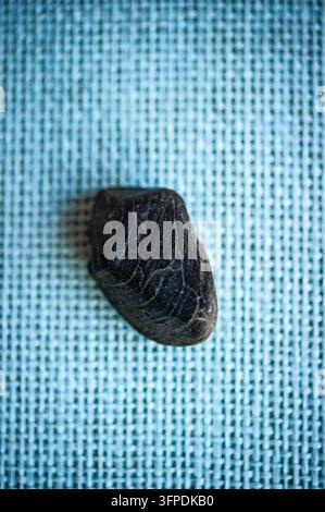 Dark textured pebble on a pale blue woven surface, photographed from above with strong contrast and visible fabric detail Stock Photo