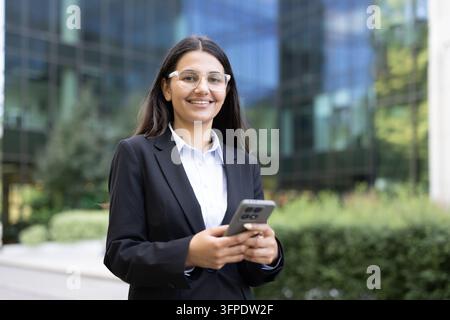 Professional businesswoman in a suit and glasses smiling at the camera while holding a mobile phone in her hands, standing outdoors in front of a modern office building Stock Photo