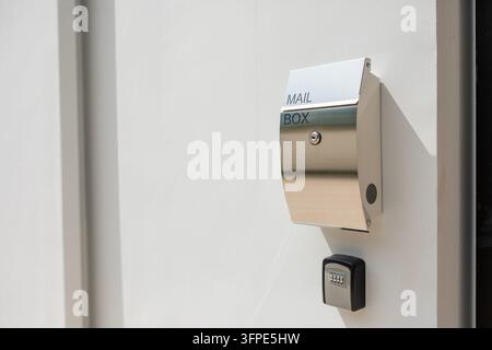 Modern stainless steel mailbox with MAIL BOX lettering mounted on white wall, keyhole lock and combination lockbox below, sunlight and shadow, large c Stock Photo