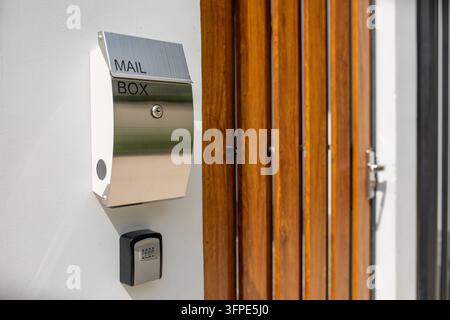 Modern stainless steel mailbox with MAIL BOX lettering on white wall beside wooden front door, keyhole lock and combination lockbox below, vertical sl Stock Photo