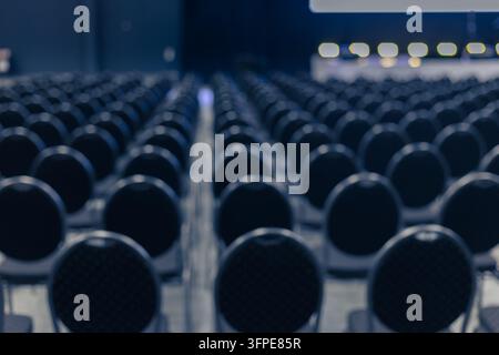 Blurred conference chairs in indoor event space form soft defocused rows leading toward stage lighting. Out of focus dark seats create elegant bokeh. Stock Photo