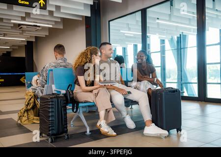 A group of three people sits together at an airport terminal with their bags. They are talking and looking relaxed. Stock Photo