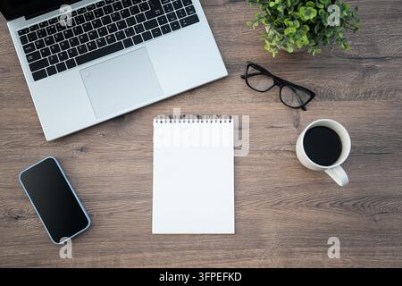Top view of home office workspace with laptop, blank notebook, smartphone, coffee cup, eyeglasses and green plant on wooden desk. White blank screens Stock Photo
