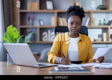 African american woman working in a home office, holding a document and writing on paper, using a laptop and calculator, focused on managing financial tasks or studying personal accounting Stock Photo