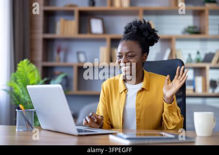Happy african woman smiling and waving during laptop video conference at her home office desk, engaging in remote work, online meeting, and virtual collaboration with confidence Stock Photo