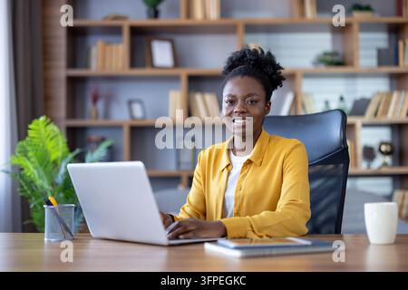 Woman working remotely from her home office, confidently smiling at the camera while using a laptop, showing professional dedication and successful digital entrepreneurship Stock Photo