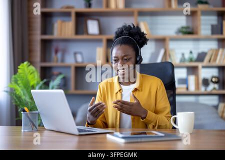 Young african american businesswoman smiling and gesturing while participating in an online meeting with a headset and laptop from her home office, representing remote work and virtual communication Stock Photo