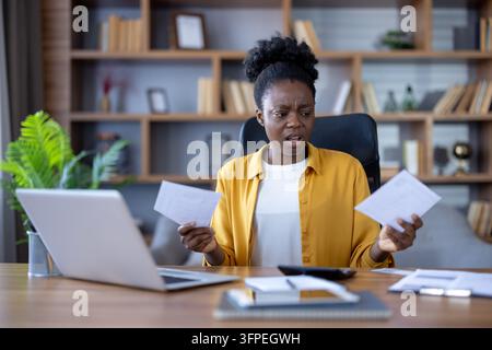 Black woman receiving shocking news, holding two bills, looking confused and stressed while managing personal finances and paperwork at her home office desk Stock Photo