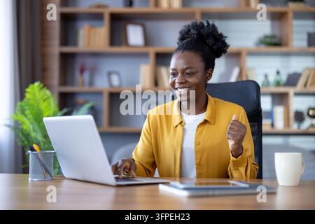 Young woman in a home office smiles and raises a fist while working at a laptop, celebrating remote work success, confidence and professional achievement on a bright desk Stock Photo