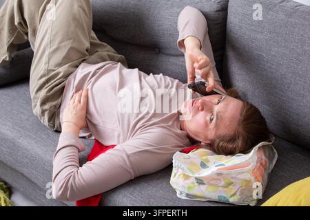 A woman lies on a cozy couch, using her phone to have a conversation. She appears relaxed, enjoying a casual moment at home in a comfortable setting. Stock Photo