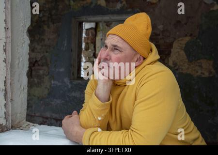 A man in a yellow hat and sweater sits by the window in an old abandoned building. He looks thoughtfully at the snowy landscape, Stock Photo