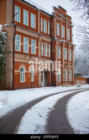 An old brick building with many windows stands in the snow next to a winding path. Trees and winter are visible in the background. Stock Photo