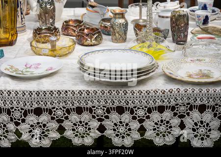 Vintage tableware displayed at a flea market, featuring floral porcelain plates, glassware, vases and bowls arranged on an elegant lace tablecloth. Stock Photo