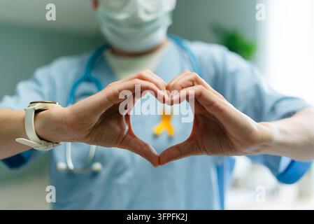 Compassionate doctor and caring nurse form heart shape with hands to show love and support for children in hospital setting. Yellow ribbon on uniform highlights awareness of pediatric cancer Stock Photo