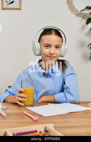 A preteen girl in headphones while holding glass of orange juice in a cozy workspace Stock Photo