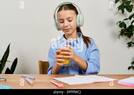 At home, a preteen girl uses her headphones for study, sipping a drink in her cozy space Stock Photo