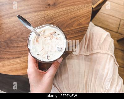 Woman holding iced latte with metal straw in glass, top view. Cold coffee drink with milk foam on wooden table. Cafe lifestyle, morning beverage. Symb Stock Photo