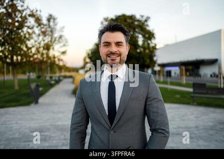 A man is smiling and wearing a suit while standing in a park. The sun is setting behind trees. Stock Photo