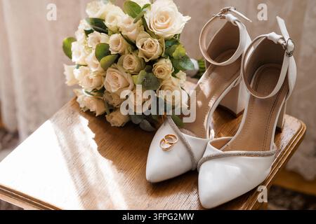 Elegant white high heel wedding shoes with gold bands adorned with sparkly trim, two wedding rings resting on shoe tip, lush bouquet of creamy white r Stock Photo