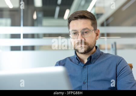 Man with glasses and beard looking intently at a laptop screen, focusing on his work in a contemporary office environment, representing dedication and professionalism Stock Photo