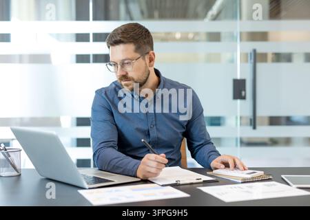 Professional man working on a laptop. Calculating figures with a calculator. And writing notes on documents. Concentrating on finance management and business strategy in a contemporary office setting Stock Photo