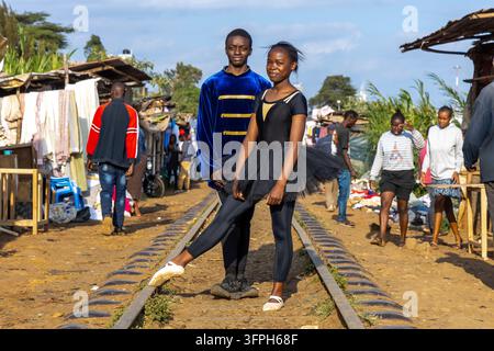 Young man in blue velvet jacket and ballerina in black tutu posing on railway tracks at the Kibera ballet school with passers-by and market stalls beh Stock Photo