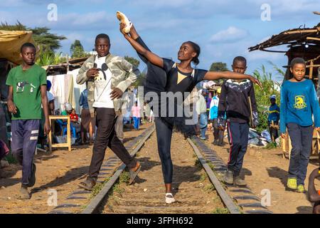 Ballerina in black tutu raising one leg in a high extension on railway tracks at the Kibera ballet school while four boys walk past and watch, Nairobi Stock Photo