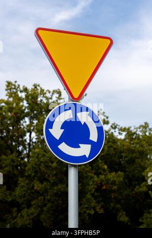 Yield and roundabout traffic signs stand mounted on metal post beside green trees and sky. Stock Photo