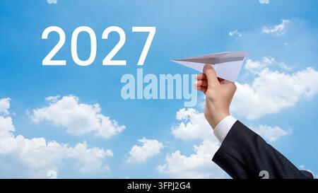 Businessman hand launching a paper airplane towards 2027 text in a blue sky. Stock Photo