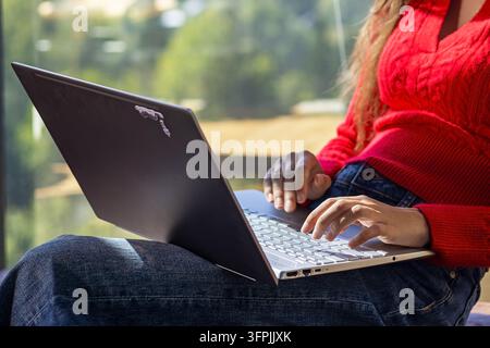 Young woman typing on laptop while sitting by a large window, close-up view of remote work and modern digital lifestyle in a bright natural setting. Stock Photo