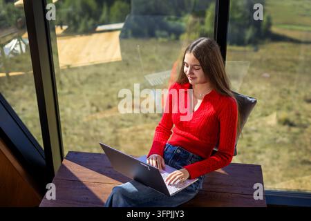 Young woman working on laptop beside panoramic window, remote work in a modern mountain cabin with peaceful countryside view and bright sunlight. Stock Photo