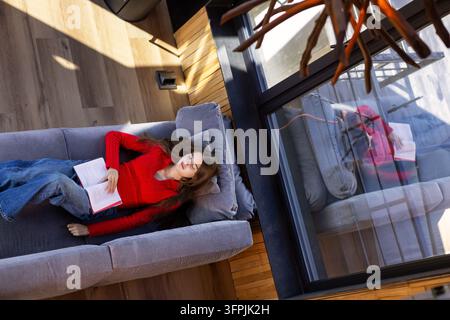 Young woman in red sweater lying on gray sofa with open book and closed eyes in modern cabin, peaceful relaxation and rest beside panoramic window in Stock Photo