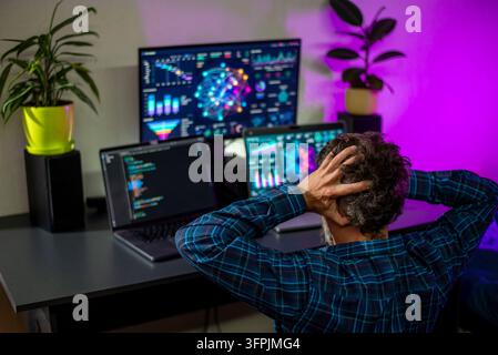Over-the-shoulder view of a developer holding his head at a desk with laptops and monitors showing code and colorful analytics dashboards. Stock Photo