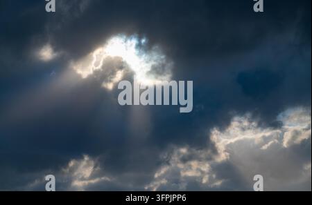 Dramatic cloudscape with bright sunlight breaking through heavy dark clouds and visible sun rays, creating a powerful atmospheric scene. Stock Photo