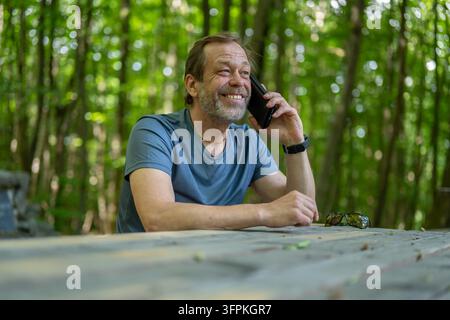 A smiling middle aged man sits at a wooden table and talks on a smartphone in a forest. Stock Photo
