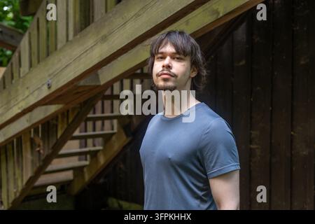 A young man stands beside wooden stairs in an outdoor setting with a calm expression. Stock Photo