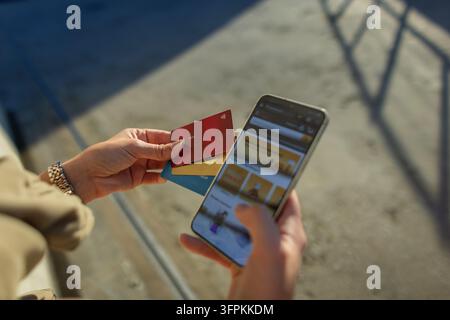 Close-up of a woman's hands holding a smartphone with an online store website open and several credit cards outdoors. Selecting a bank card for a secu Stock Photo