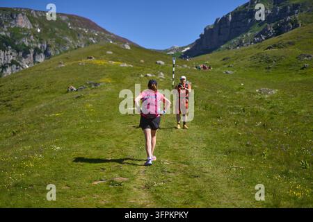 Woman with pink backpack walking uphill on a grassy trail while a man films her approach in an alpine valley in Bucegi mountains, Romania Stock Photo