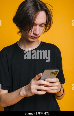 Young man using smartphone with both hands against bright yellow studio background wearing black t-shirt and simple jewelry focused neutral expression Stock Photo
