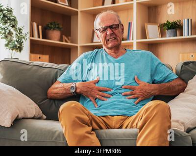 Aged man grimacing from abdominal cramps and digestive problems while resting on couch, experiencing discomfort and illness. Stock Photo