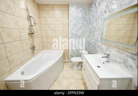Modern bathroom interior with corner bathtub, sink vanity and toilet. Bright washroom with beige wall tiles, abstract accent wall, large mirror and clean contemporary design Stock Photo