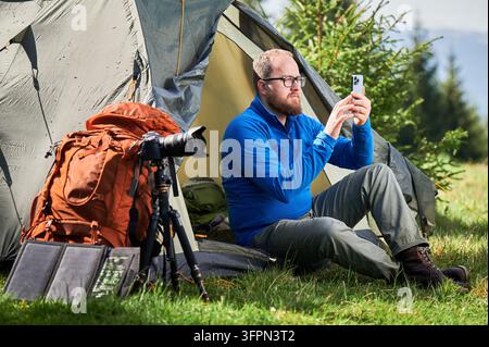 Camper uses smartphone beside tent with camera gear, backpack, and portable solar panel in mountain campsite. Useful for outdoor travel, mobile technology, remote work, and adventure marketing Stock Photo