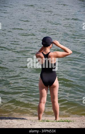 Woman adjusting goggles before open water swimming. Female swimmer in black swimsuit and cap standing by lake, back view, preparing for training, summer outdoor activity Stock Photo