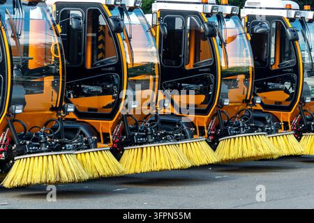 Several orange street sweepers aligned in a row, with large brushes visible in the foreground, parked on an asphalt surface Stock Photo