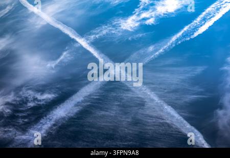 Crossing contrails create a bold saltire against a vivid blue sky, surrounded by wispy cirrus clouds and soft light, evoking motion, aviation, contras Stock Photo